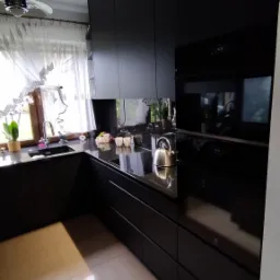 Nowoczesna, czarna kuchnia z błyszczącymi frontami, czarnym blatem z granitowym wzorem, srebrnym czajnikiem i beżowymi płytkami podłogowymi. W tle okno z firanką i lampa sufitowa.