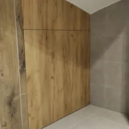 Ukryte drzwi w ścianie wykończonej drewnopodobnymi panelami, zintegrowane z aranżacją łazienki, na tle szarych płytek i jasnej podłogi.