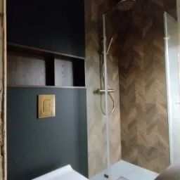 Nowoczesna łazienka z kabiną prysznicową, drewnianą mozaiką na ścianach, podwieszaną toaletą i ciemnymi szafkami z drewnianymi p&oacute;łkami. Złota armatura prysznicowa i przycisk spłuczki.