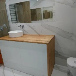 Nowoczesna łazienka z białą umywalką nablatową na drewnianym blacie, białe szafki pod umywalką, lustro w ramie pasującej do blatu, białe płytki imitujące marmur.