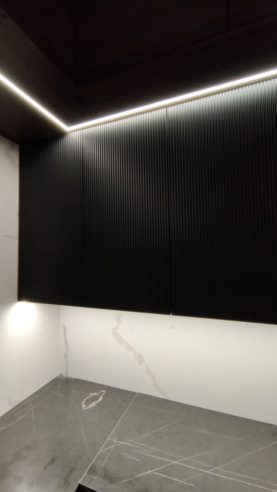 Czarna ściana z ryflowanymi panelami podświetlona listwą LED u góry i dołem, marmurowe płytki na ścianach i podłodze.