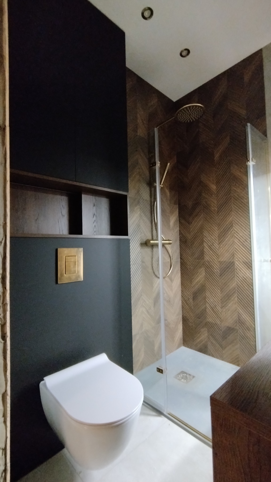 Nowoczesna łazienka z kabiną prysznicową, drewnianą mozaiką na ścianach, podwieszaną toaletą i ciemnymi szafkami z drewnianymi półkami. Złota armatura prysznicowa i przycisk spłuczki.