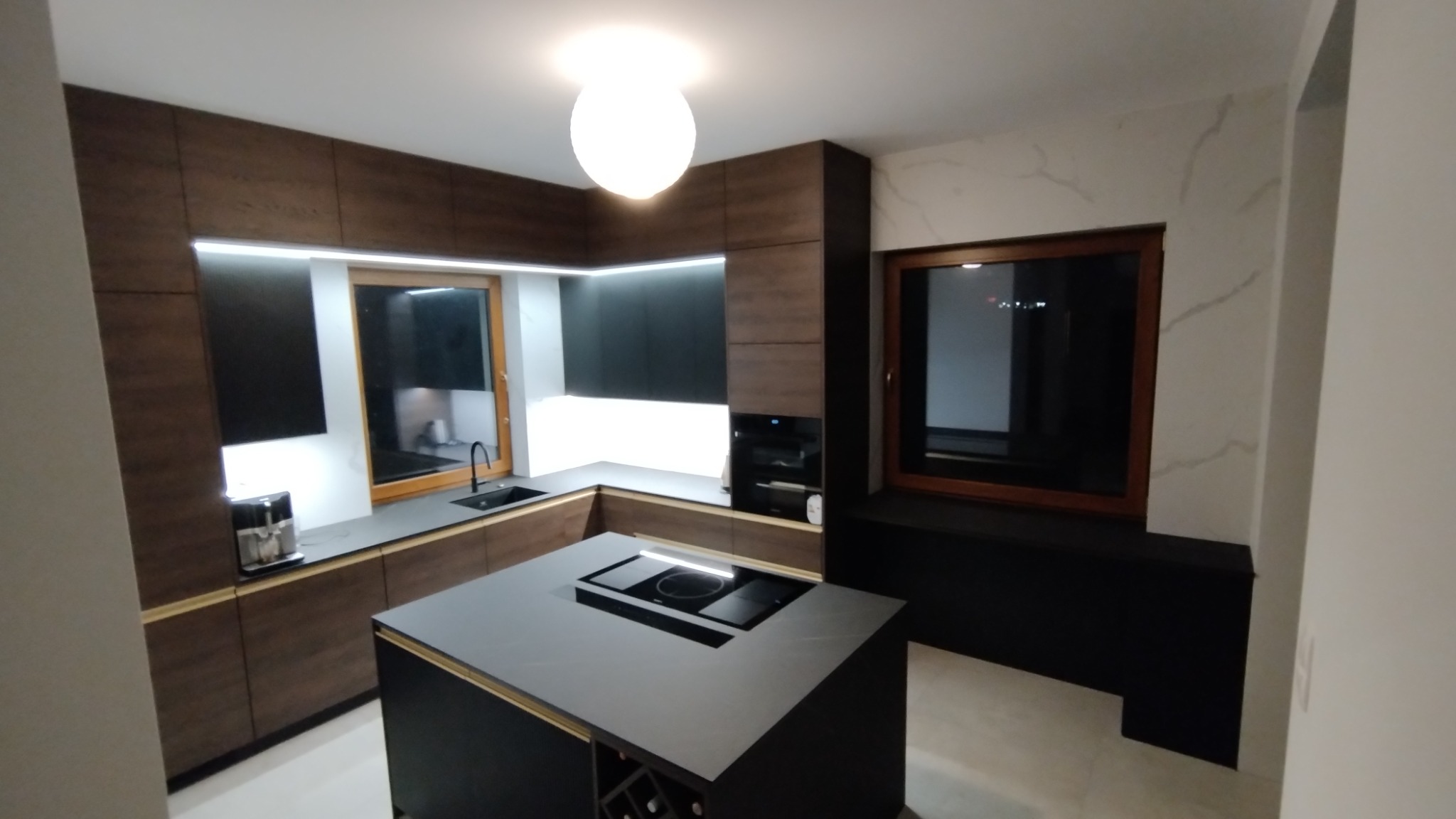 Nowoczesna kuchnia z drewnianymi frontami, czarnymi blatami i wyspą kuchenną z płytą indukcyjną, oświetlona lampą sufitową i taśmami LED pod szafkami.