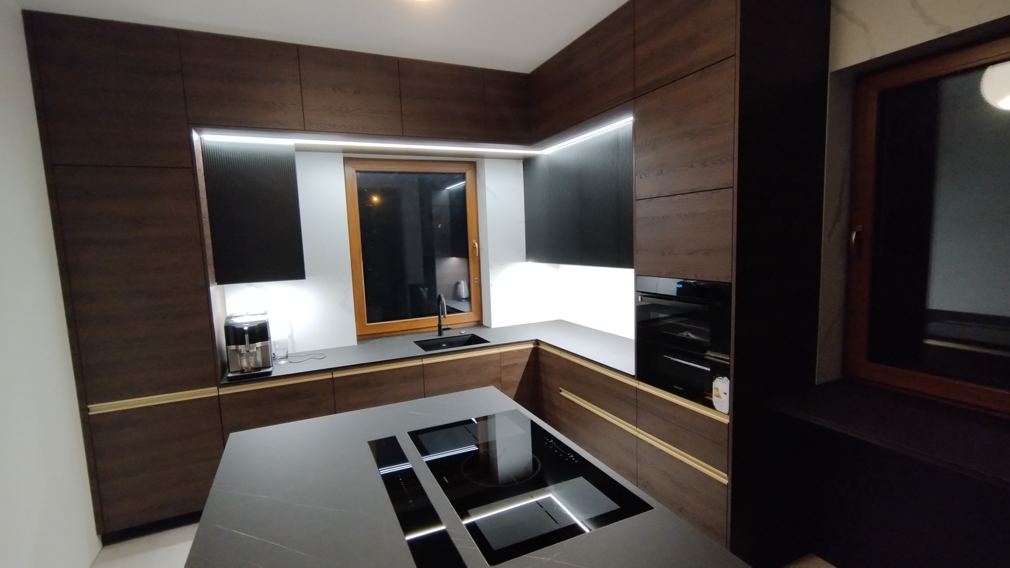 Nowoczesna kuchnia z ciemnego drewna z wyspą kuchenną i oświetleniem LED, widok na okno i ekspres do kawy.