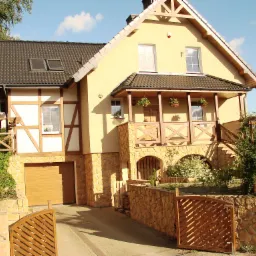 Dwukondygnacyjny dom jednorodzinny z jasną elewacją, elementami muru pruskiego i kamienną podmurówką, otoczony zielenią, z drewnianą bramą wjazdową i garażem z bramą segmentową.