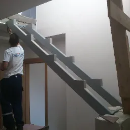 Montaż szarej, nowoczesnej konstrukcji schodów w surowym wnętrzu; widoczny pracownik w koszulce z logo firmy, wskazujący na górną część schodów.