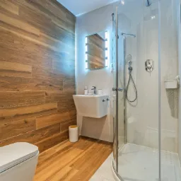 Nowoczesna łazienka z drewnopodobnymi płytkami na ścianie i podłodze, podwieszana umywalka z podświetlanym lustrem, kabina prysznicowa z przezroczystego szkła i biała toaleta z rolką papieru...