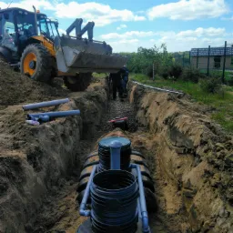 Instalacja przydomowej oczyszczalni ścieków w wykopie; widoczny ciągnik z łyżką, rury kanalizacyjne i pracownicy.