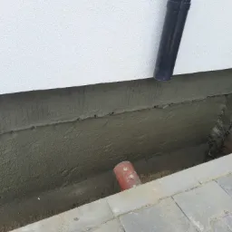 Świeżo nałożona warstwa izolacyjna na fundamencie budynku, widoczny fragment rury spustowej i rura w wykopie, obok ułożona kostka brukowa.