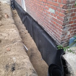 Akma Polska Sp.z.o.o. - Czarne folie kubełkowe zabezpieczają fundament ceglanego budynku przed wilgocią. W wykopie widoczny pracownik z łopatą i rolka folii. Prace ziemne w toku.