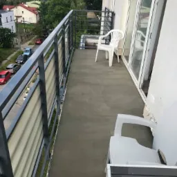 Wykończony balkon z szarą, betonową posadzką, metalową balustradą z wypełnieniem z blachy falistej i dwoma białymi krzesłami z tworzywa, widok na okoliczne budynki i zieleń.