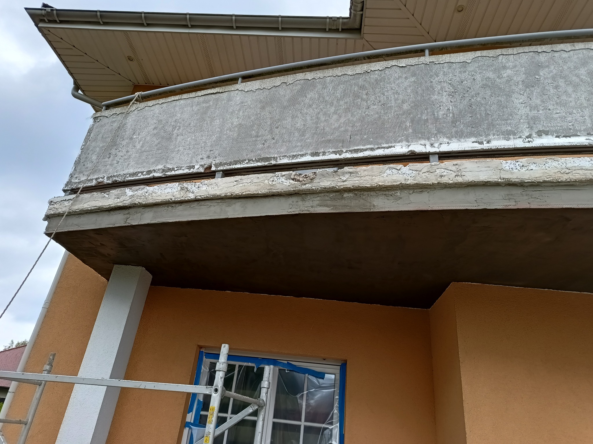 Balkon w trakcie remontu z widocznymi śladami starej farby i nową warstwą szpachli, okno zabezpieczone folią malarską, drabina oparta o ścianę budynku.