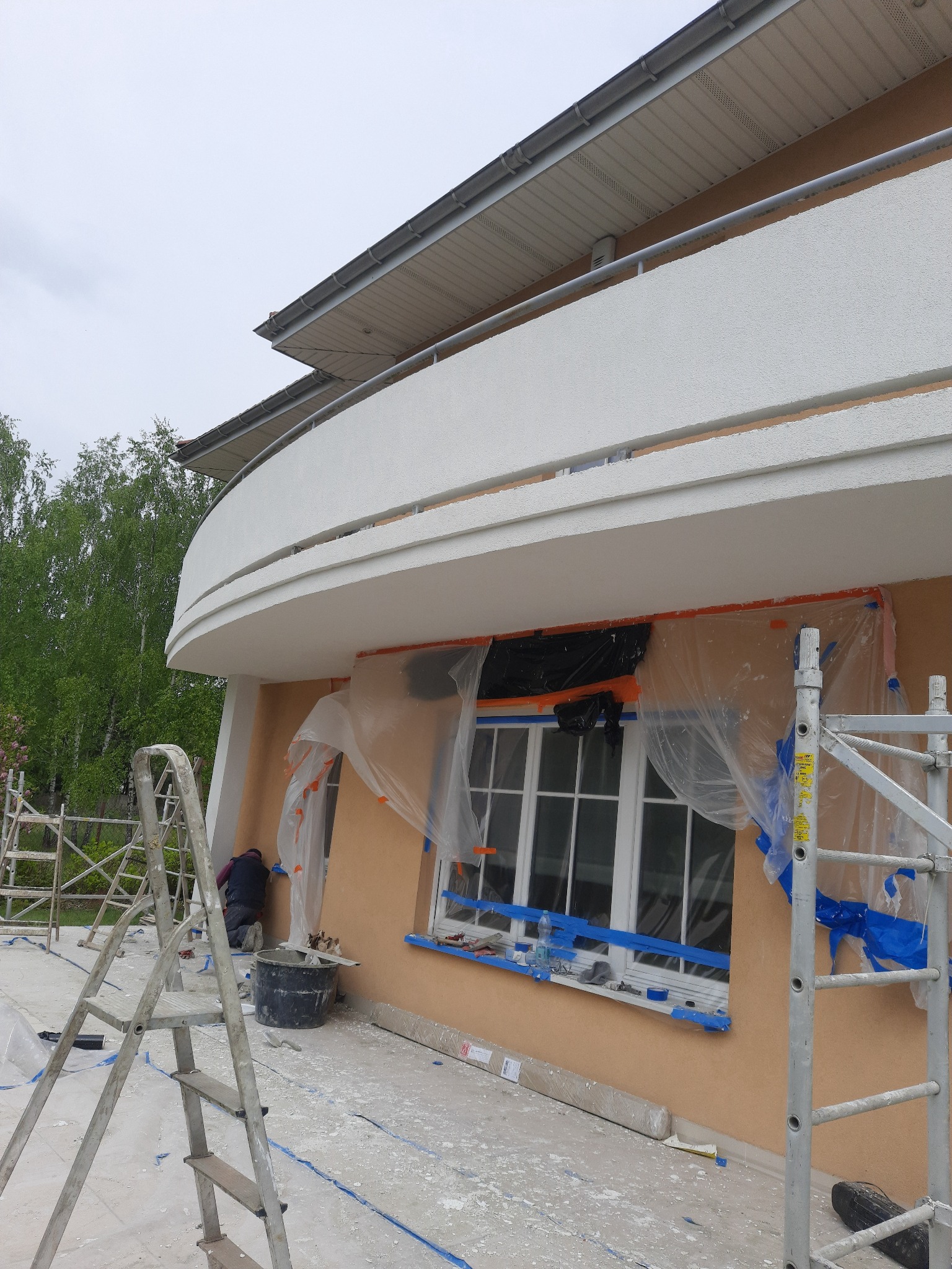 Prace wykończeniowe elewacji budynku z zaokrąglonym balkonem, okna zabezpieczone folią, widoczne rusztowania i pracownik przy oknie.