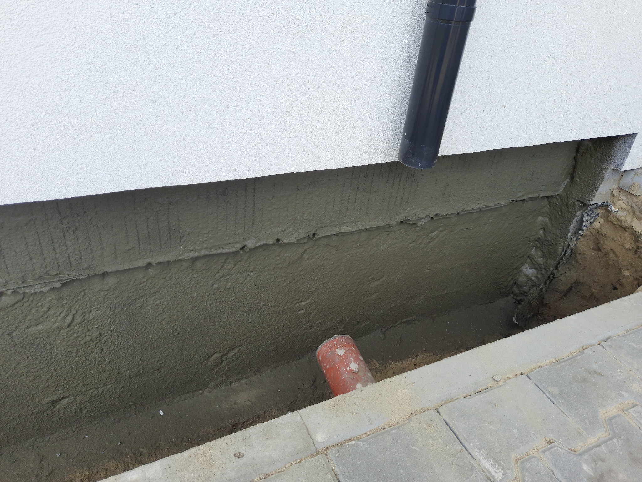 Świeżo nałożona warstwa izolacyjna na fundamencie budynku, widoczny fragment rury spustowej i rura w wykopie, obok ułożona kostka brukowa.