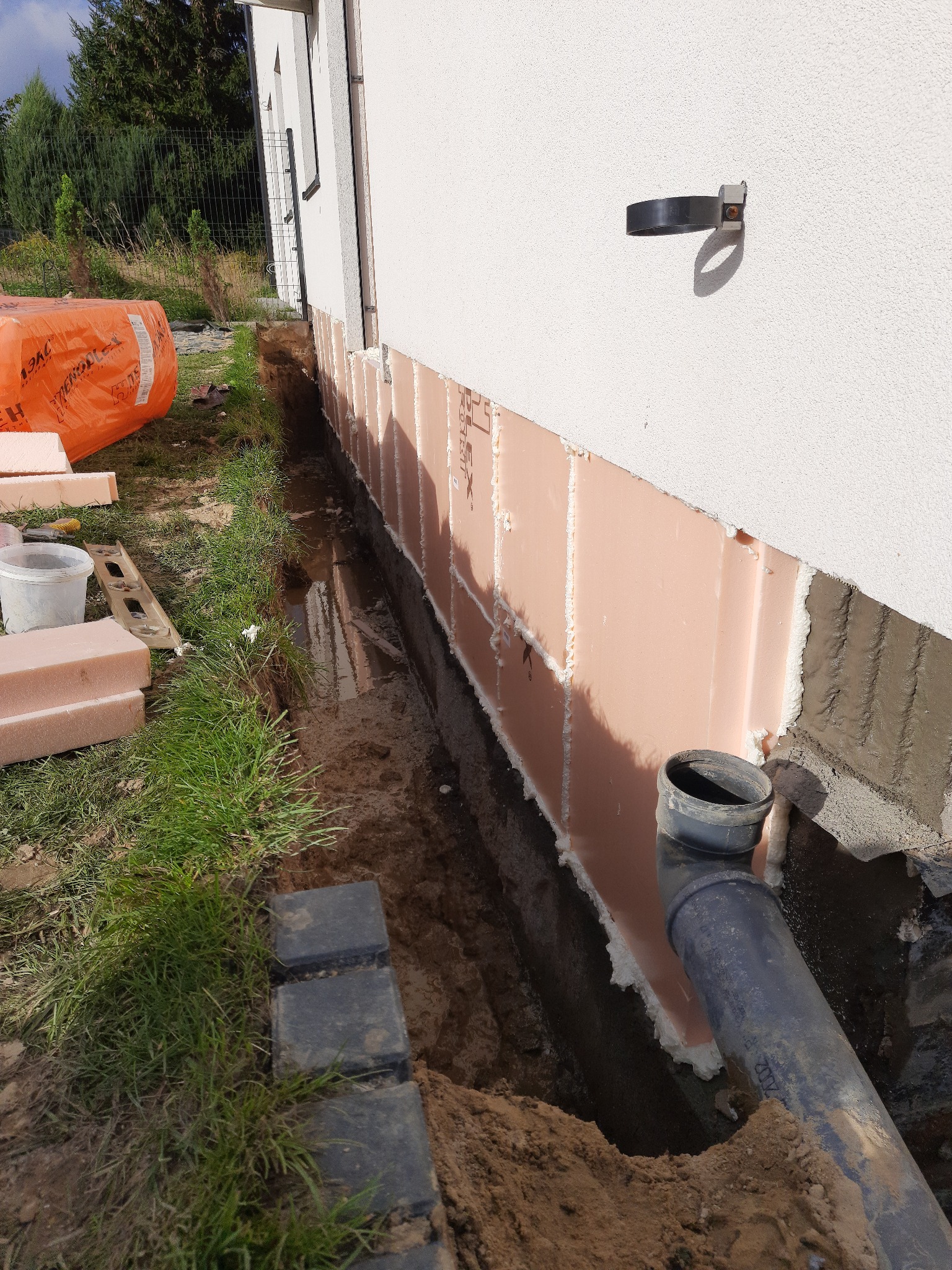 Ocieplenie fundamentów budynku jednorodzinnego z użyciem różowych płyt izolacyjnych, widoczny wykop wzdłuż ściany i rura kanalizacyjna.