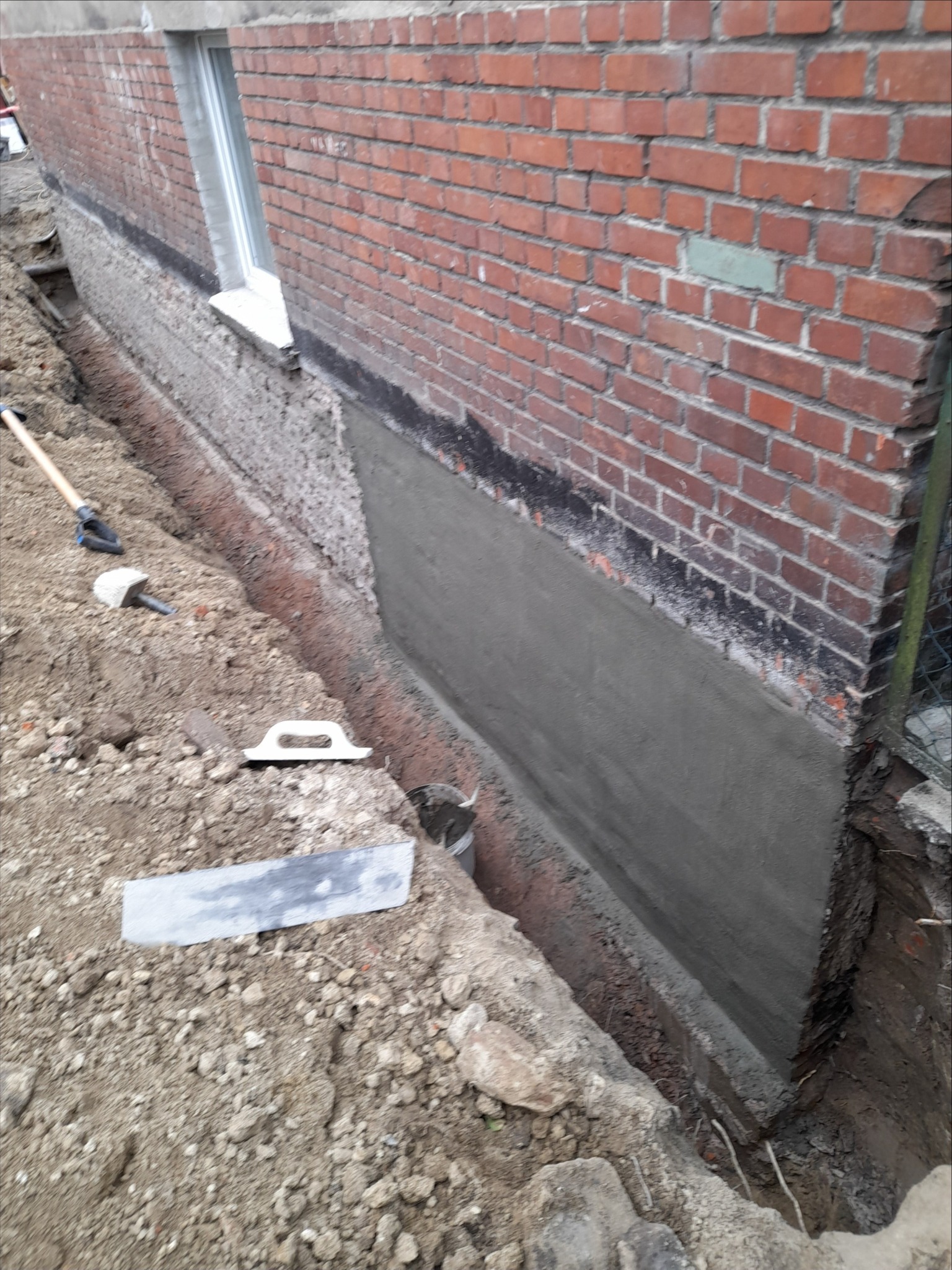 Ceglany fundament budynku z nałożoną warstwą szarego tynku, widoczne narzędzia murarskie i wykop. Prace ziemne w toku, fragment okna w tle.