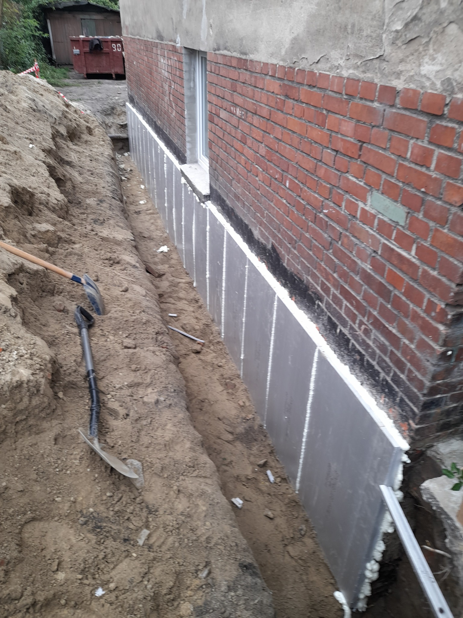 Odkopane fundamenty budynku z cegły, zabezpieczone szarymi płytami izolacyjnymi i pianką montażową. W wykopie leżą dwie łopaty. Widoczny kontener na gruz.