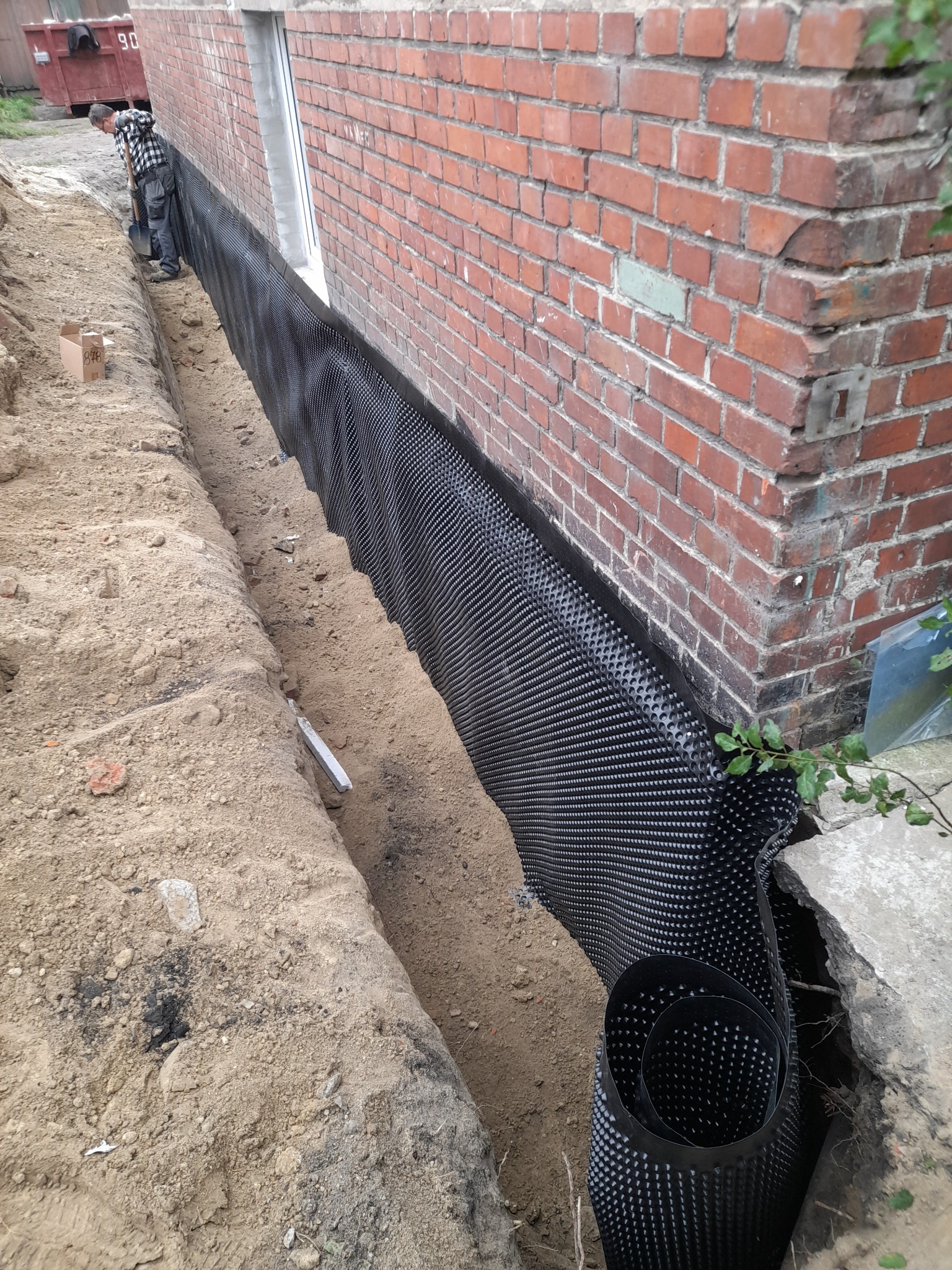 Czarne folie kubełkowe zabezpieczają fundament ceglanego budynku przed wilgocią. W wykopie widoczny pracownik z łopatą i rolka folii. Prace ziemne w toku.