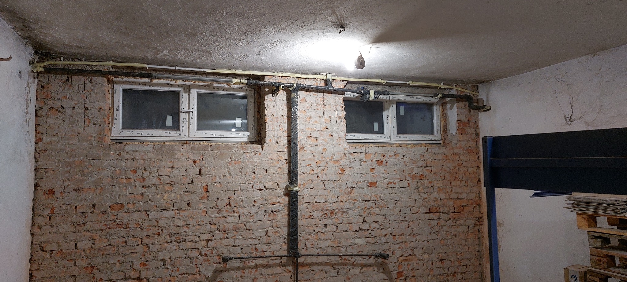 Surowa ściana z cegły z dwoma oknami PCV, rurami i przewodami. Widoczne ślady po instalacjach. W tle fragment regału i palety. Nierówna powierzchnia sufitu.