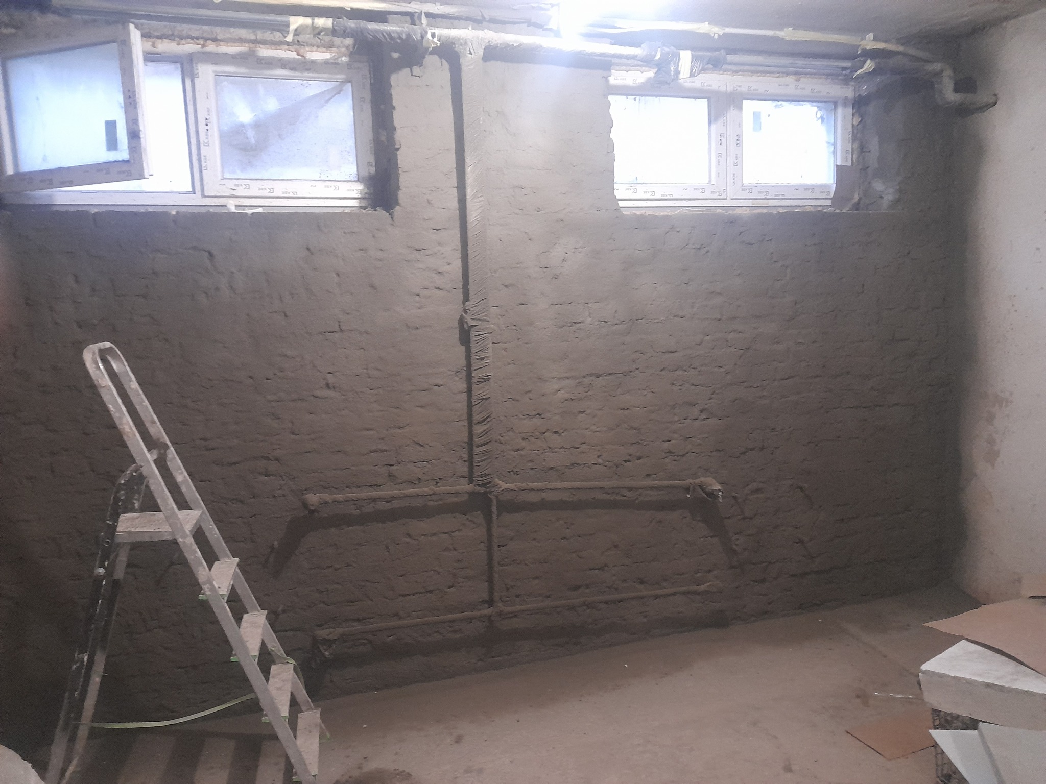 Otynkowana ściana piwnicy z dwoma oknami, widoczne rury i drabina. Prace wykończeniowe w surowym wnętrzu. Szare tło, brak dekoracji.