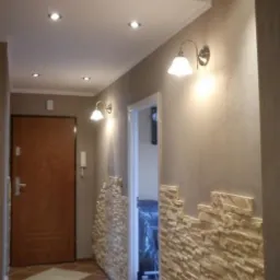 Korytarz po remoncie z dekoracyjną ścianą z kamienia, brązową podłogą w karo i oświetleniem punktowym oraz kinkietami.