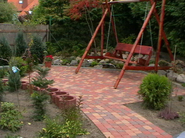 Ułożona z czerwono-brązowej kostki brukowej ścieżka i taras ogrodowy z drewnianą huśtawką w tle, obrzeża z elementów betonowych i nasadzenia roślinne.
