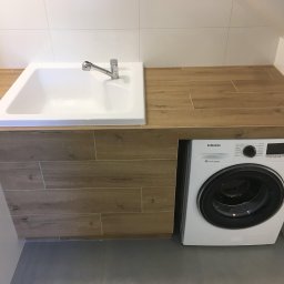 Mat-Gaw - Nowoczesna łazienka z białą umywalką wpuszczoną w drewnopodobny blat, pod którym znajduje się pralka Samsung, całość wykończona białymi płytkami.