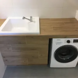 Nowoczesna łazienka z białą umywalką wpuszczoną w drewnopodobny blat, pod którym znajduje się pralka Samsung, całość wykończona białymi płytkami.