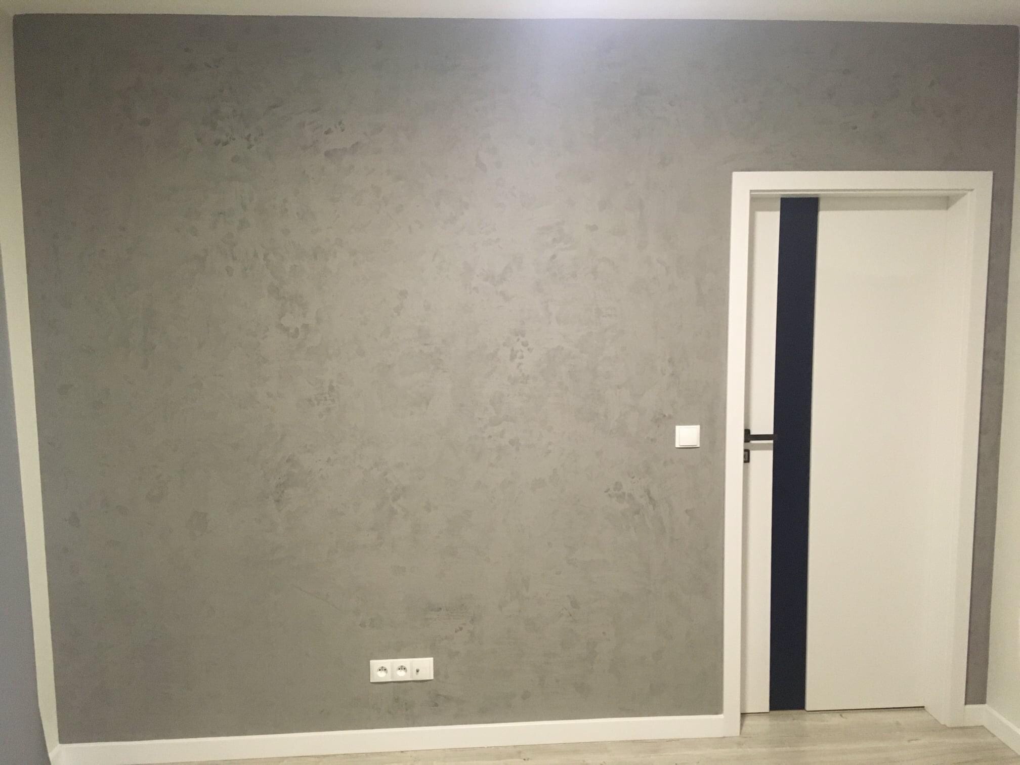 Szara ściana z efektem dekoracyjnym, białe listwy przypodłogowe i ościeżnica, białe drzwi z pionowym, ciemnym pasem, gniazdka elektryczne i włącznik światła.
