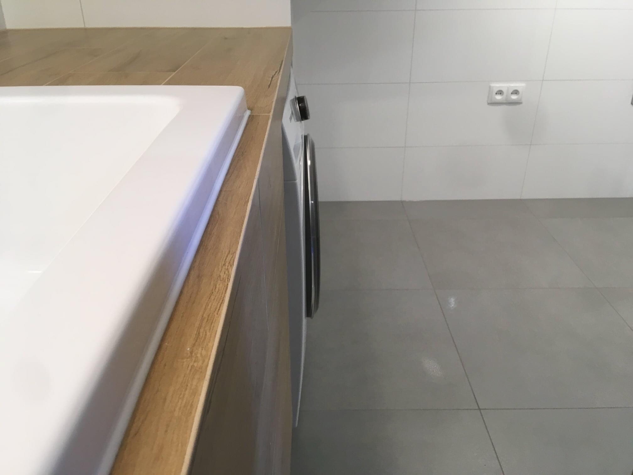 Fragment wykończonej łazienki z blatem imitującym drewno, białą umywalką i pralką, widok na płytki ścienne i podłogowe w odcieniach bieli i szarości.