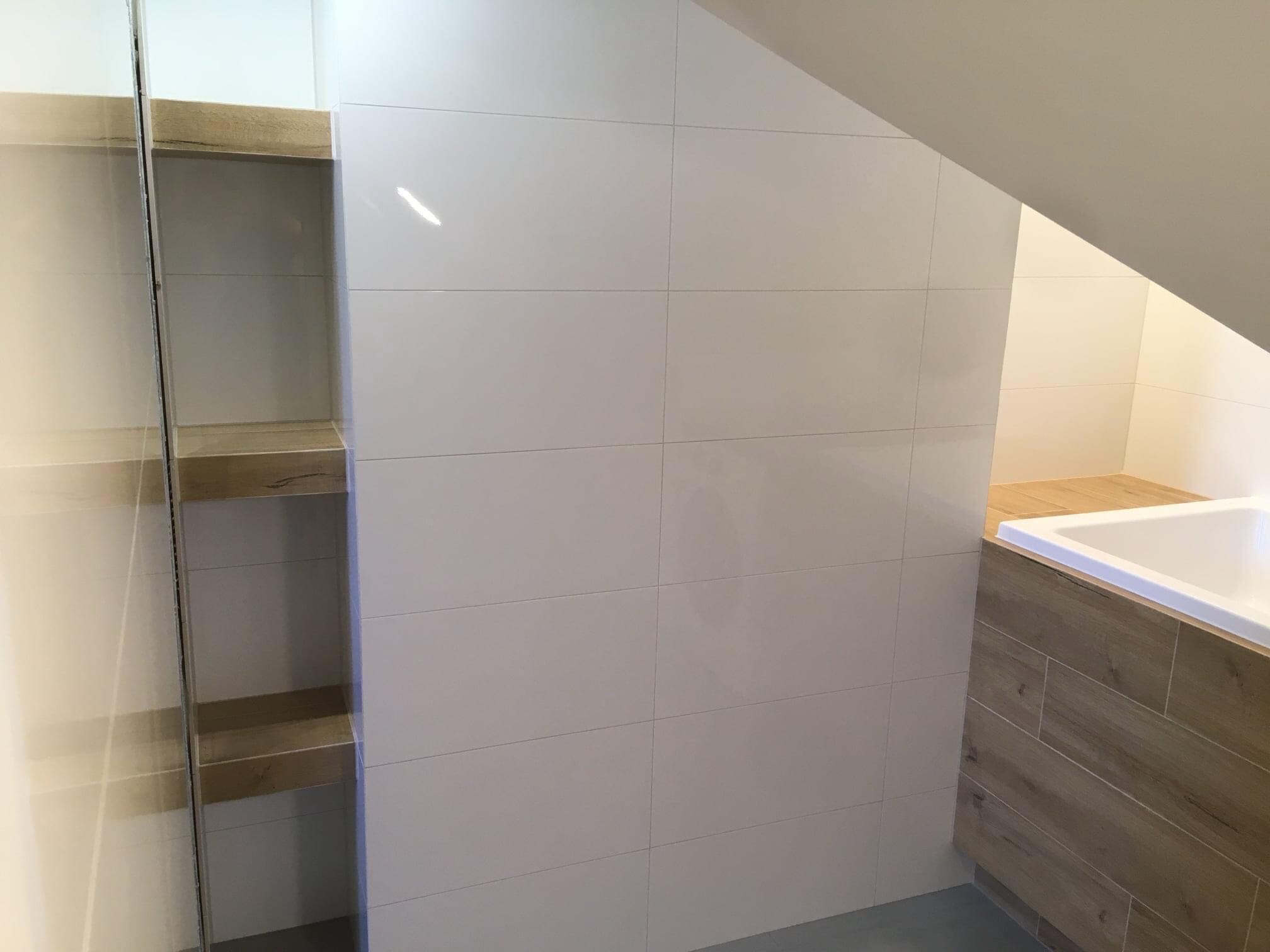 Nowoczesna łazienka na poddaszu z białą glazurą, obudową wanny imitującą drewno i półkami wnękowymi z drewnianymi elementami.