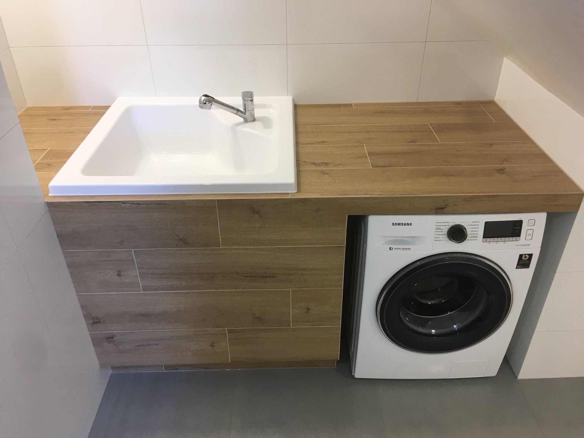 Nowoczesna łazienka z białą umywalką wpuszczoną w drewnopodobny blat, pod którym znajduje się pralka Samsung, całość wykończona białymi płytkami.