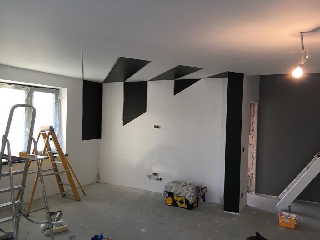 Wnętrze pokoju w trakcie malowania z geometrycznymi wzorami w kolorze czarnym na suficie i ścianach, widoczne drabiny malarskie i narzędzia pracy.