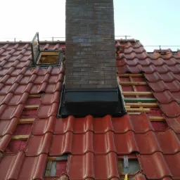 Czerwona dachówka karpiówka z oknem dachowym i kominem z cegły podczas prac naprawczych, widoczne elementy więźby i izolacji.