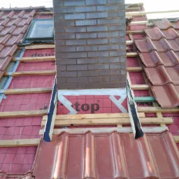 Montaż ceglanego komina na dachu pokrytym czerwoną dachówką, widoczne elementy konstrukcyjne, membrana dachowa z napisem 'top' oraz okno dachowe w tle.