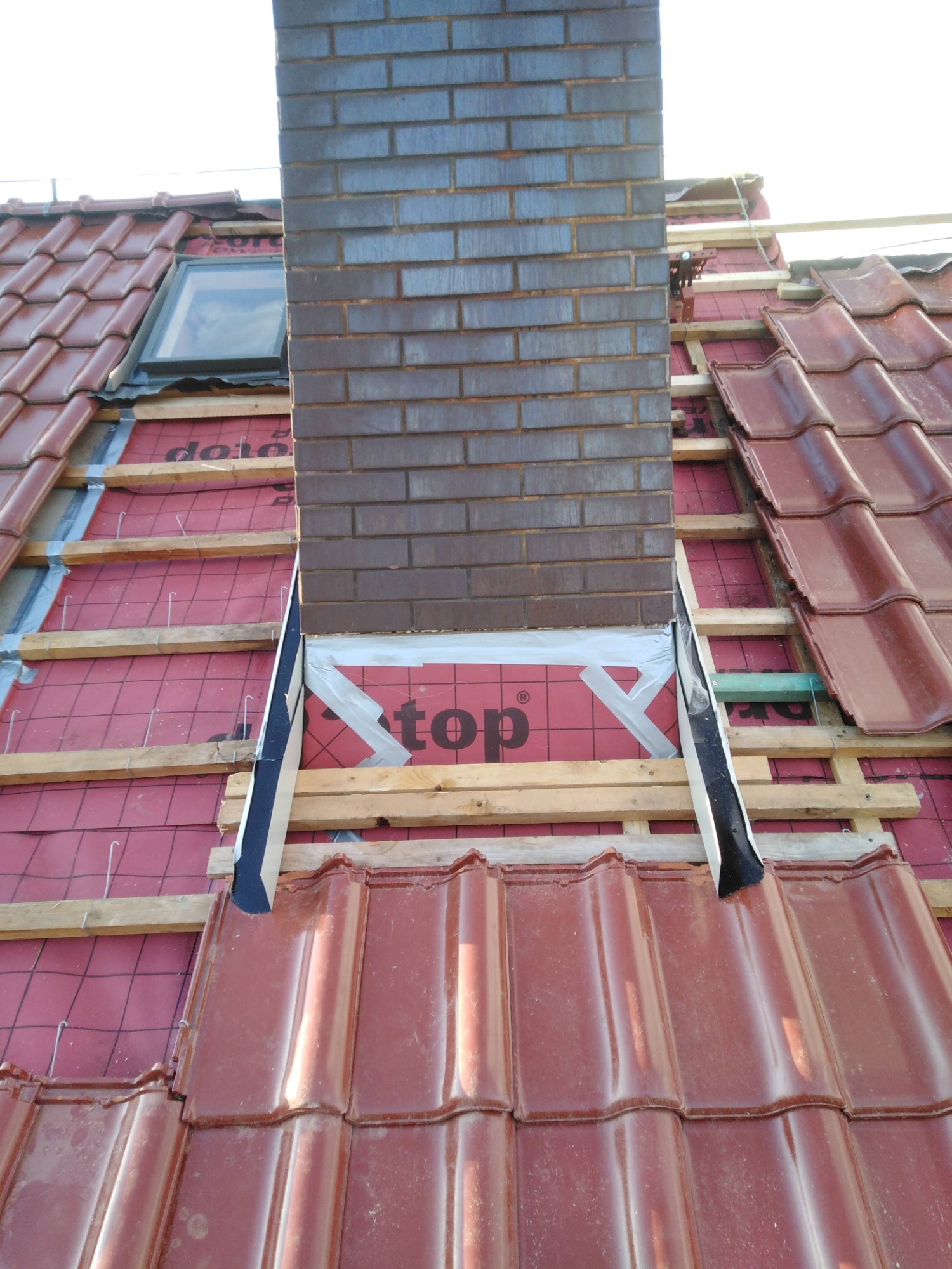 Montaż ceglanego komina na dachu pokrytym czerwoną dachówką, widoczne elementy konstrukcyjne, membrana dachowa z napisem 'top' oraz okno dachowe w tle.