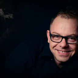 Portret uśmiechniętego mężczyzny w okularach na ciemnym tle, z delikatnym zarysem klasycznego aparatu Nikon w tle.