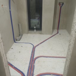 sanitec - Podłogowe rury grzewcze w trakcie montażu, układane w wylewce, widoczne rury w kolorze czerwonym i niebieskim, pion kanalizacyjny i butelka ze spryskiwaczem przy oknie.
