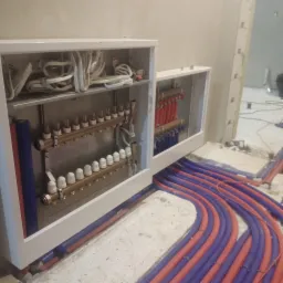 Rozdzielacz ogrzewania podłogowego w białej szafce, widoczne rury w kolorze czerwonym i niebieskim, surowy stan posadzki.