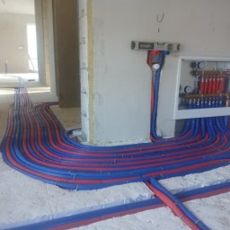 sanitec - Rozłożone rury ogrzewania podłogowego w kolorze niebieskim i czerwonym, przymocowane do styropianu, z rozdzielaczem na ścianie w tle i poziomicą. Widoczne surowe ściany i otwór drzwiowy.
