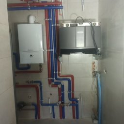 F.U.H Sanitec - Biały piec gazowy Vaillant i szary piec zamontowane na ścianie z płytkami. Widoczne rury w kolorze niebieskim i czerwonym. Instalacja grzewcza w kotłowni.