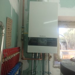 F.U.H Sanitec - Biały piec gazowy Buderus zamontowany na ścianie z widocznymi rurami miedzianymi i stalowymi. W tle okno i rozdzielacz instalacji grzewczej.