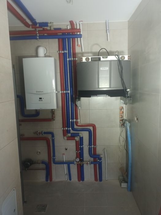 Nowa instalacja grzewcza z piecem gazowym Vaillant i szarym zasobnikiem w pomieszczeniu wyłożonym jasnymi płytkami, widoczne rury w kolorach niebieskim i czerwonym.