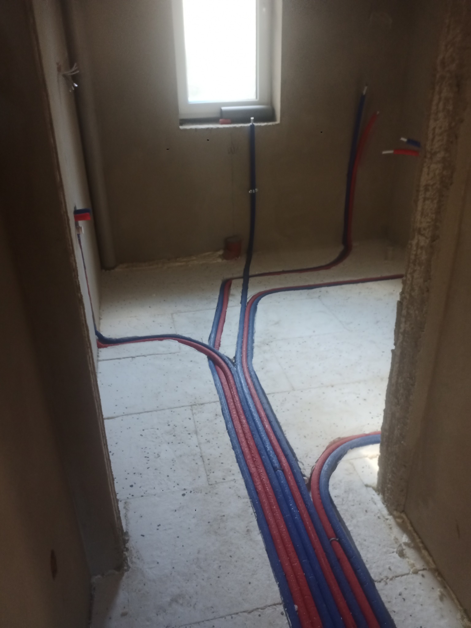 Instalacja ogrzewania podłogowego w surowym stanie w Skarszewach: widoczne rury grzewcze czerwone i niebieskie, zalane w wylewce, prowadzone pod oknem i wzdłuż ścian, przed dalszym wykończeniem.