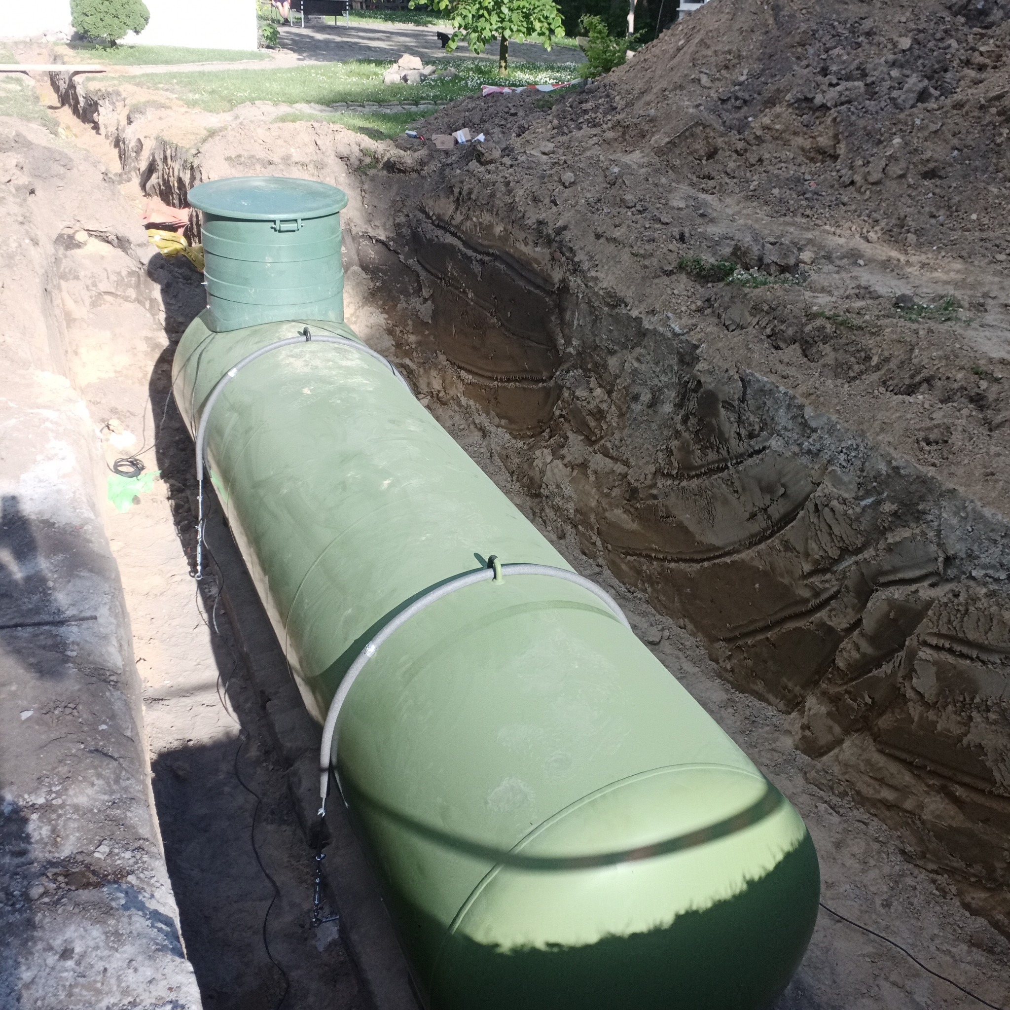 Zielony zbiornik zakopany w wykopie ziemnym, widoczne ślady po koparce na ścianach wykopu, z boku leżą kable.