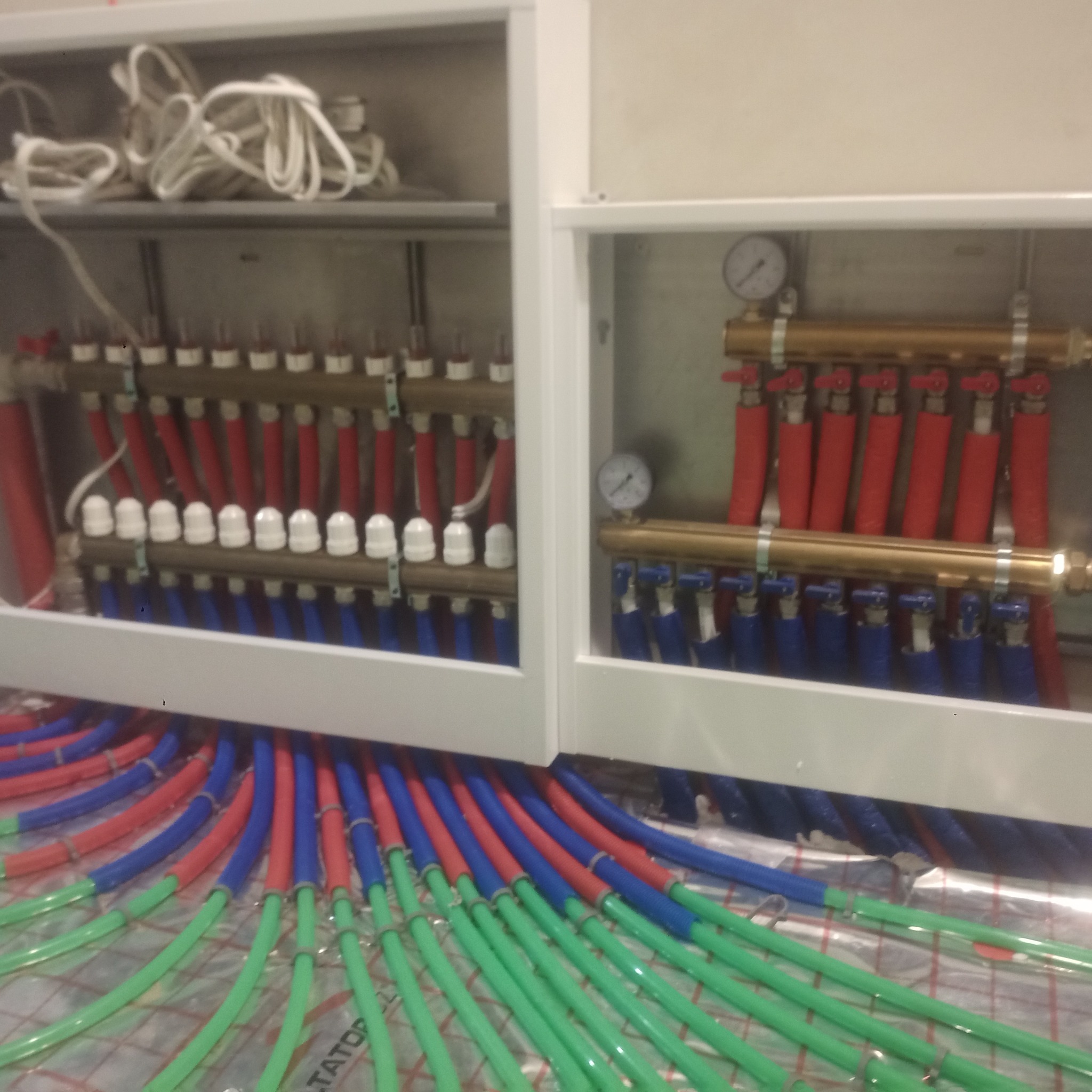 Rozdzielacz ogrzewania podłogowego w szafce, widoczne rury w kolorach zielonym, niebieskim i czerwonym oraz manometry.