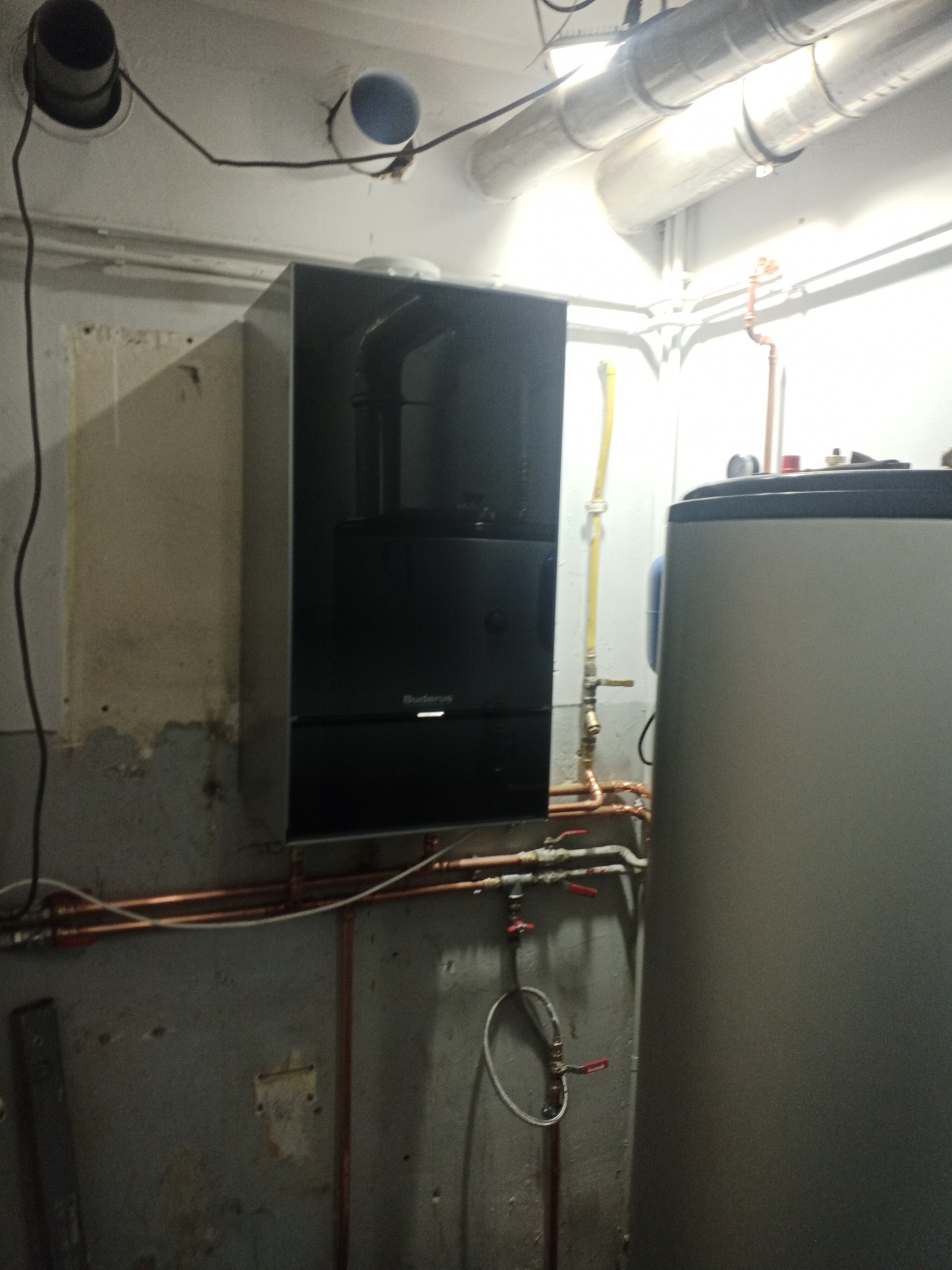 Czarny piec gazowy Buderus zamontowany na szarej, surowej ścianie piwnicy, widoczne rury miedziane i zawory, obok zasobnik ciepłej wody, oświetlenie jarzeniowe na suficie.