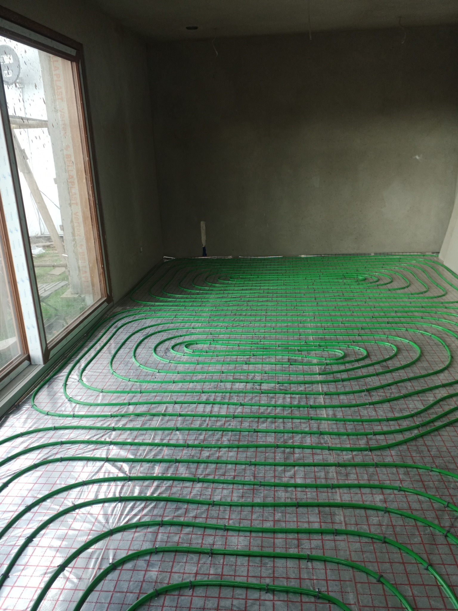 Zielone rurki ogrzewania podłogowego ułożone spiralnie na folii z czerwoną siatką, widoczne okno z kroplami deszczu i surowe ściany w szarym pomieszczeniu.