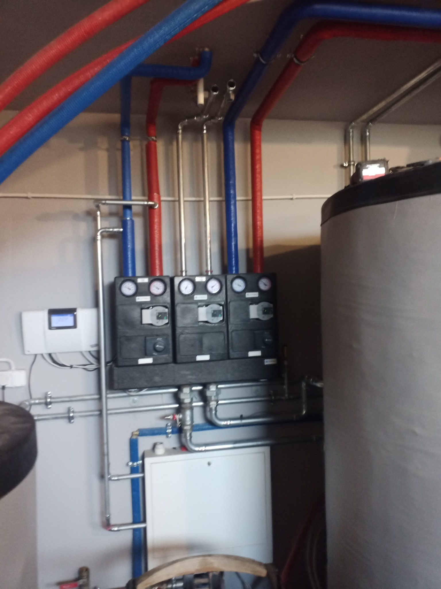 Rozbudowana instalacja grzewcza z trzema czarnymi panelami sterującymi z manometrami, zbiornikiem akumulacyjnym i rurami w izolacji czerwonej i niebieskiej, zamontowana na szarej ścianie.