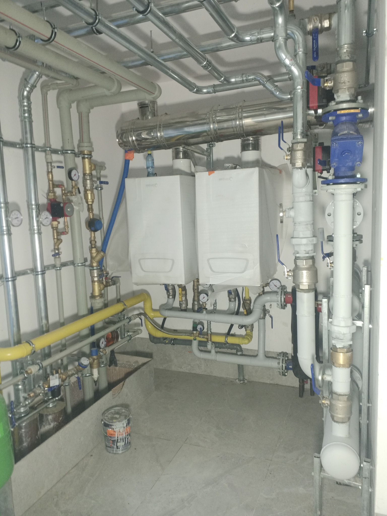 Dwa białe piece gazowe wiszące na ścianie, połączone licznymi rurami z zaworami i manometrami, z żółtą rurą gazową i farbą na podłodze.